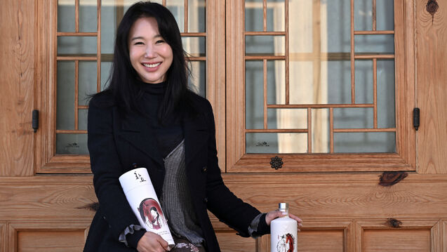 A Korean woman with long styled brown hair sits in front of a light blond wood door with traditional Korean firmaments in the window. She holds Hwayo soju bottles in her hand (resting on the out of view bench she sits on) and in her lap. 