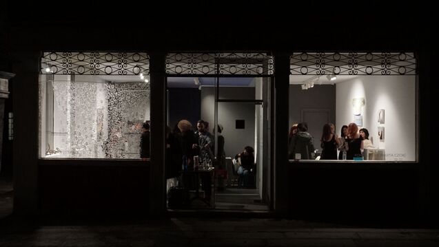 The illuminated storefront of the Czok Foundation at night, viewed from the street. Through large windows and an open door, visitors mingle inside the gallery, with Anya Lomachenko's intricate textile work Undersky visible in the left window.