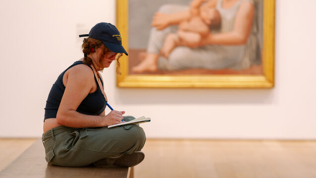 A person with light skin tone wearing a baseball hat sits on a bench with a sketchpad in front of a painting.