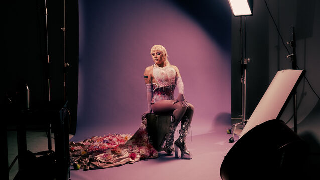 A drag performer poses in a photoshoot with a purple background