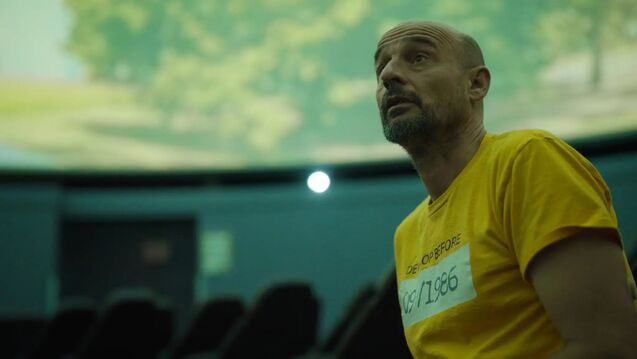  Artist Jan Tichy looks upward inside a planetarium dome, surrounded by a projected green landscape, wearing a yellow t-shirt reading "Develop Before 09/1986."
