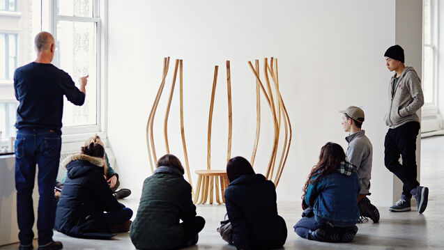 A group of students sitting in an art room looking at an art sculpture while the professor is explaining.