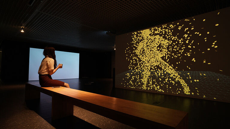 A student viewing a projected video of various yellow chips in a dark gallery room.