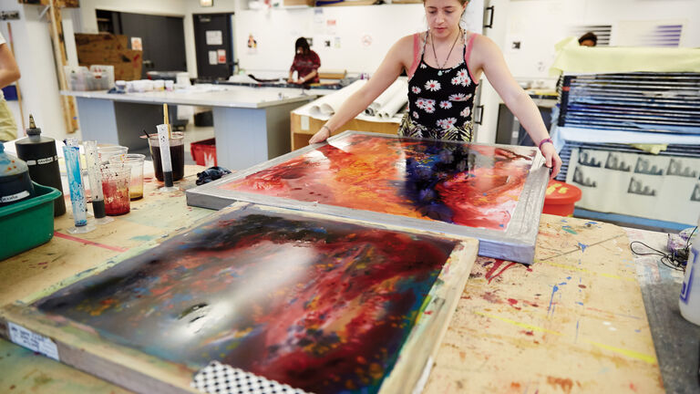 Student in foreground holding large screenprint covered in colorful ink. Other students in background working.