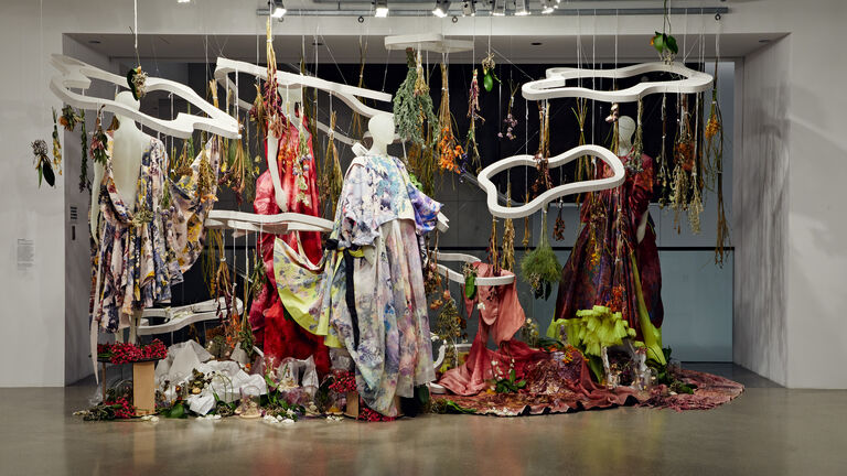 Artwork by Agnes Hamerlik. Several mannequins clothed in floral garments and surrounded by dried flowers hanging upside down and large sculptural objects suspended from the ceiling.