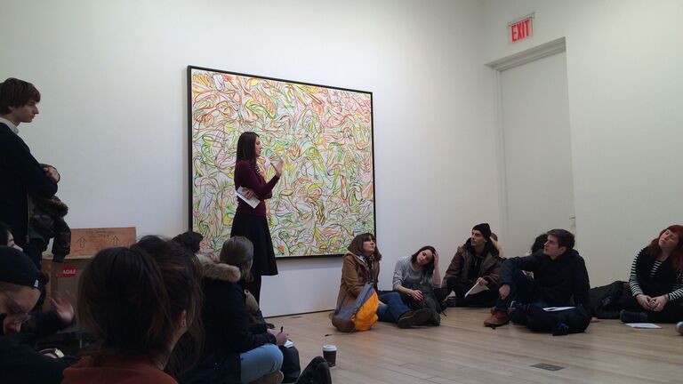 A class sits in a gallery listening to a talk.