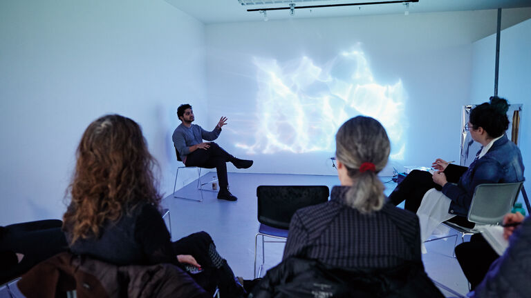 A professor sitting in a gallery with students observing a projection of a white and blue lights.