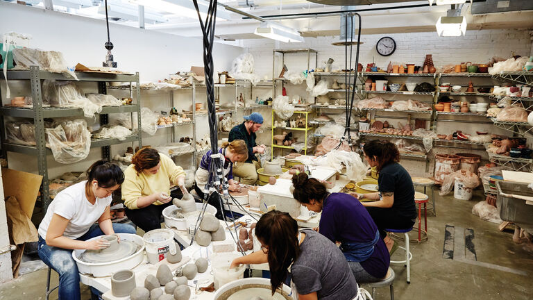 A wide shot of a ceramics studio, featuring students working with pottery wheels and other tools.