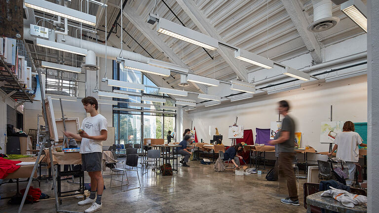 Students in a large, well-lit painting studio with a vaulted ceiling standing at easels painting on canvases.