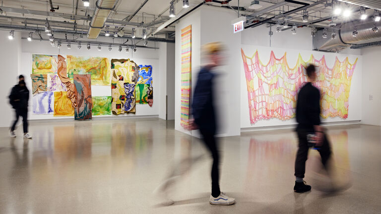 Patrons walking through galleries with bright pink and yellow textile hanging on the wall in foreground and painted textiles and canvases in background wall.