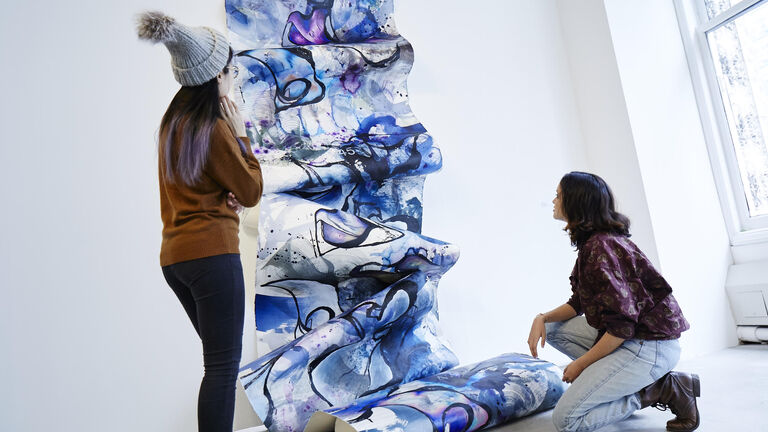Two students looking at a long piece of canvas vertically cascading down a wall covered in blues an purples and black swirls in watercolor.
