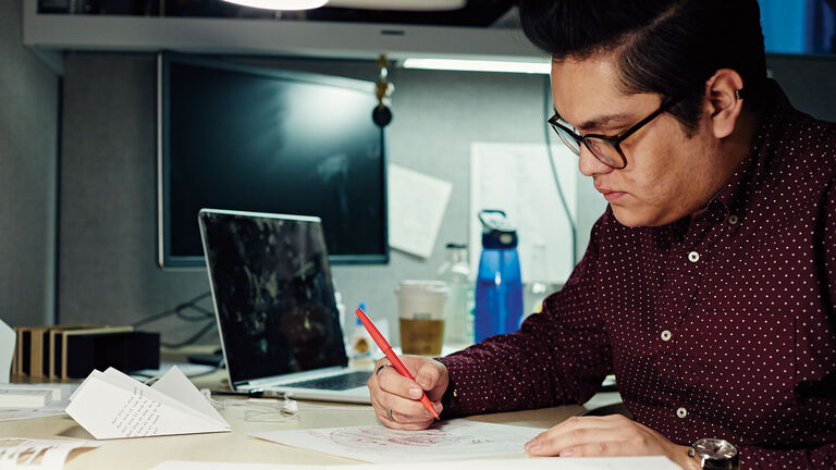 A person sitting at a desk using a red pen in front of a computer.