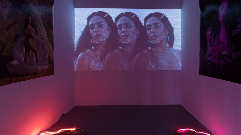 A dark room with photographs along the walls, pink neon on the floor and a video playing.