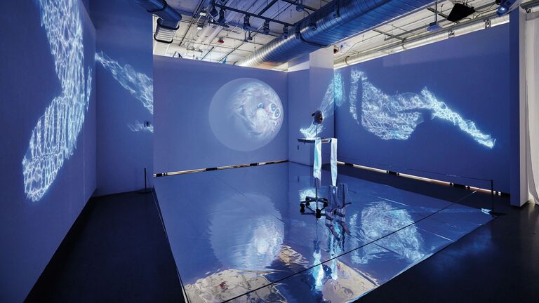 A projector placed in the center of a large mirror on the floor of a gallery with various videos being projected on the walls. 