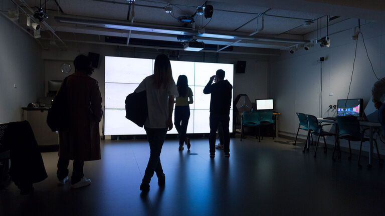 A small group of people standing in font of multiple lit up monitors. 