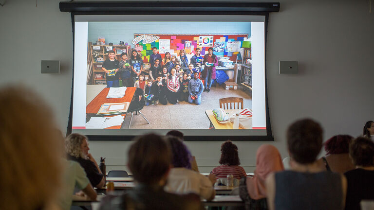 An image of a large projected screen showing a classroom of students smiling at the camera. 