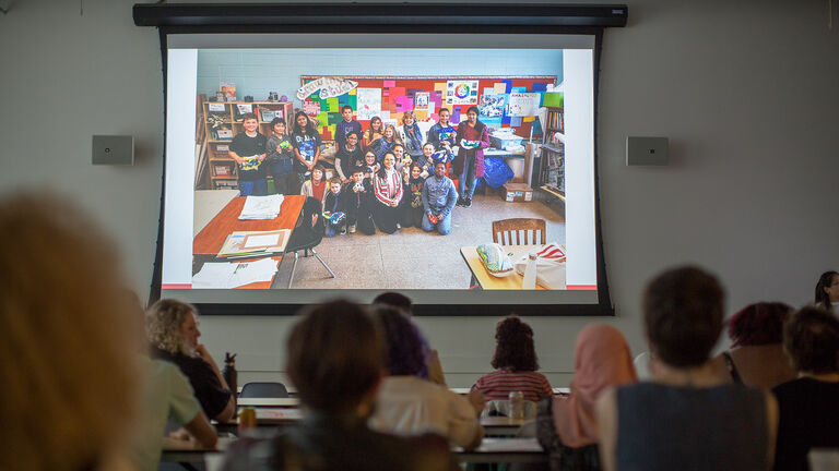 An image of a large projected screen showing a classroom of students smiling at the camera.
