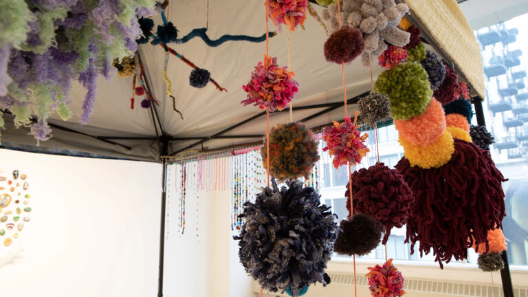 Various, colorful pompoms strung up on the frame of a tent in a gallery space.
