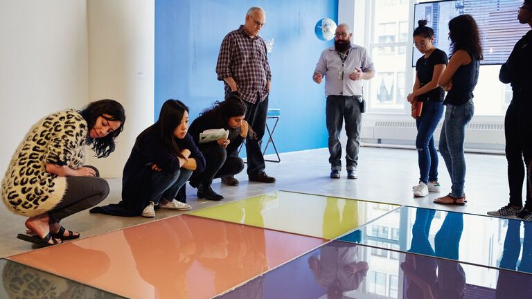 Arts Admin students listening to instructor in gallery space while looking at large, brightly colored tiles on floor.