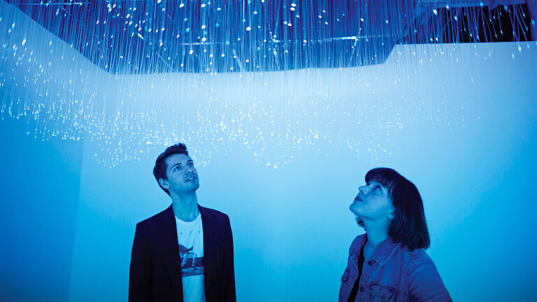 Two people looking up at a lighting installation hanging from the ceiling bathing the space in blue light.