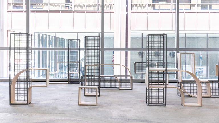 An art piece comprised of wooden furniture combined with metal grids sits in a windowed room.