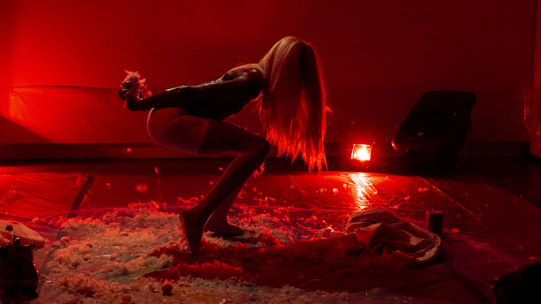 A person dances in a red-lit room with a floor of feathers