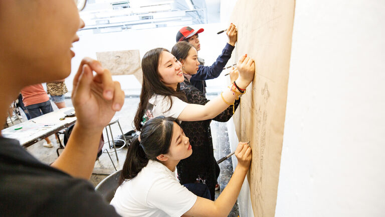 Four students writing on a piece of paper hanging on a wall.
