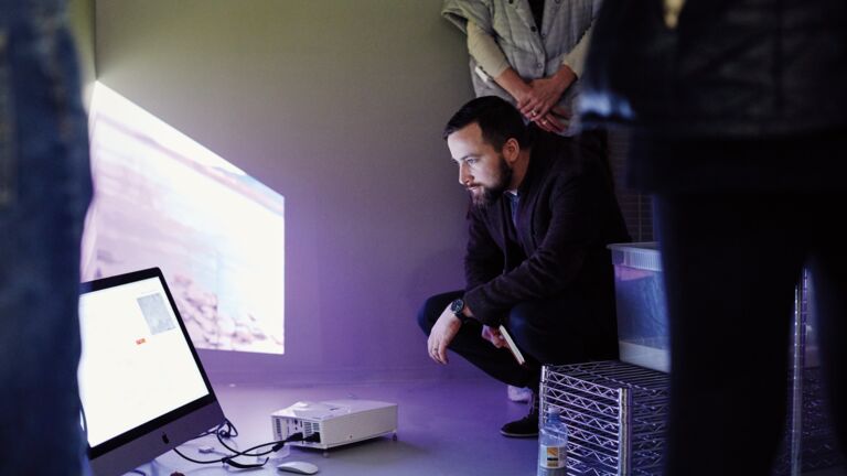 A person crotching down to look at a monitor on the floor.