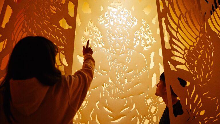 An image of two people observing an orange paper cut out piece of art with a soft white light.