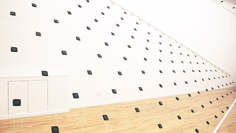 A lot of small, black microphones strung up on wires positioned in a white room with light wood floors.