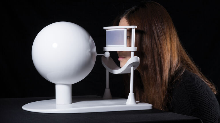 A person rests their chin on a device with a visor next to a white sphere