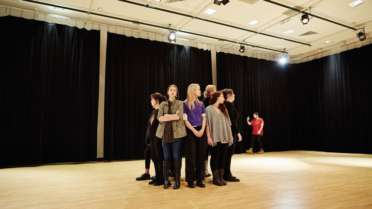 A group of students stands in a circle in a large curtained room