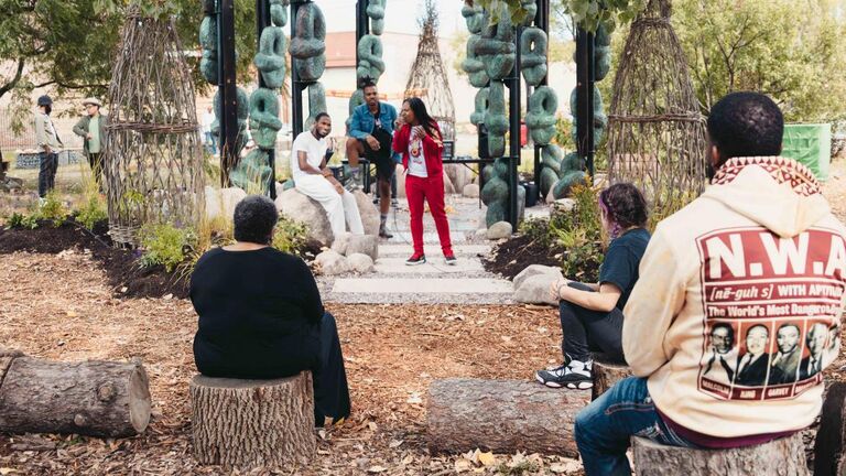 Group of people seated outside on tree stumps listening to a speaker standing under a gazebo-like structure surrounded by sculptural objects.
