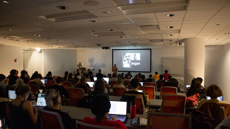 A large group of students sitting in a dark room watching a projector at the front of the room. 