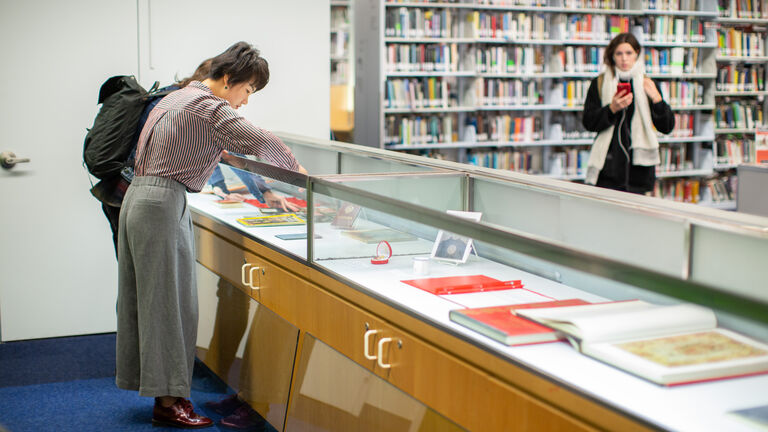 Two people looking at books in a glass case inside a library. 