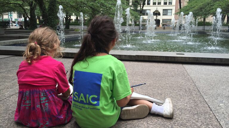 Two children sit in front of a fountain with sketchbooks.