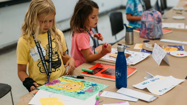 Two young students drawing pictures of people at a large craft table.