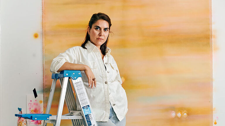 Artist Maryan Taghavi leans against a ladder next to an abstract painting