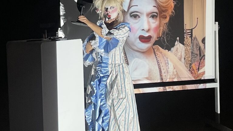 A woman in an elaborate costume stands in front of a screen, emphasizing her unique outfit and the display behind her.
