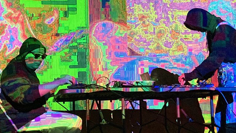 Two people at a DJ table with brightly colored lights in the background