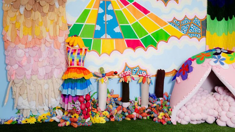 A vibrant display with colorful textiles, a rainbow dress, playful decorations, and a cozy pink hut surrounded by artificial flowers.