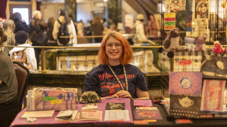 A student at the Art Sale wearing an SAIC lanyard smiling and sitting behind a table displaying various artworks.
