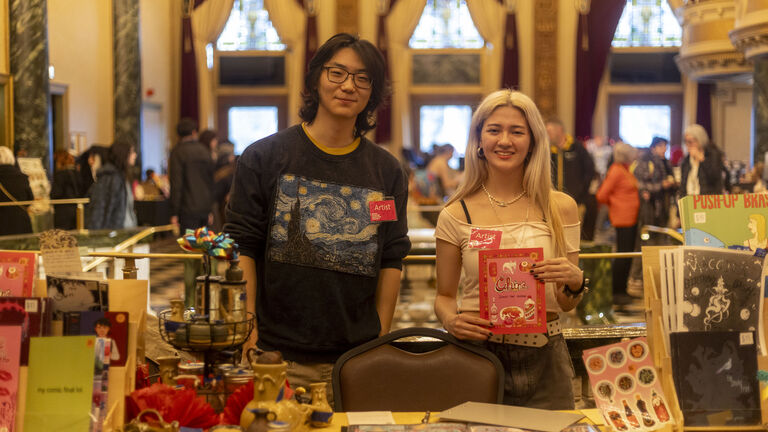 Two students at the Art Sale wearing "Artist" badges smiling and standing behind a table of various works of art.