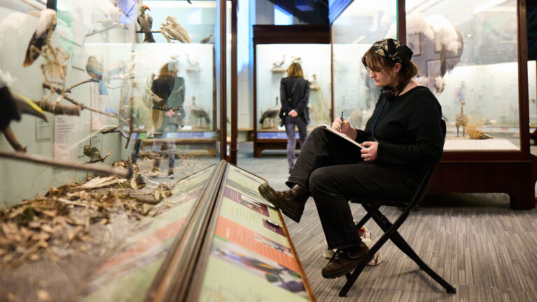 A student sits in front of a display case in a museum writing.
