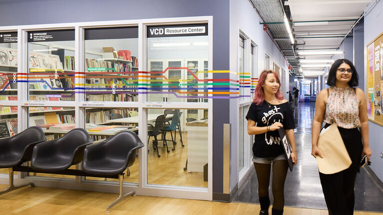 Two students walk down a hallway next to a classroom with glass windows.