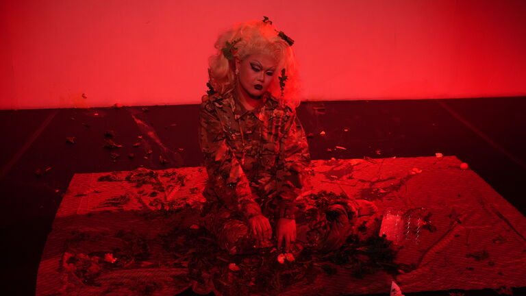 Guanqiao sits in full Drag makeup and army garments in the 2M space. 
