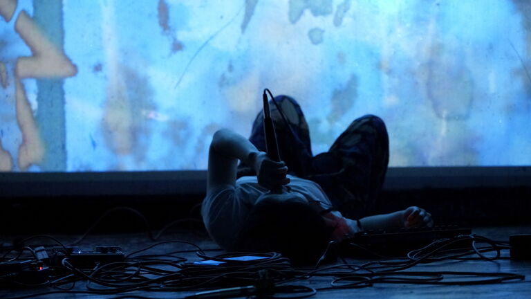 Mue is on the ground speaking into a microhpone while a visual plays in the background during a performance. 