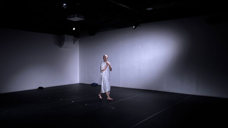 Ruby walks in dramatic lighting in the 2M space. They are wearing a hospital gown and the lighting is ghostly. 