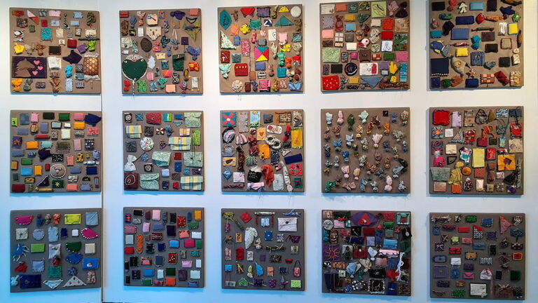 Fifteen square panels mounted on a white wall, each covered with small colorful fabric and beaded bundles arranged in grid-like patterns. Part of Sonya Clark’s The Beaded Prayers Project (1998–present).