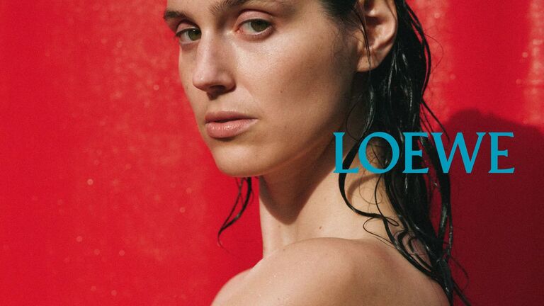Close-up portrait of actor Eva Victor shown from the shoulders up against a vivid red backdrop, with wet hair and bare shoulders, looking directly at the camera. The word “LOEWE” appears in blue type on the right side of the image.